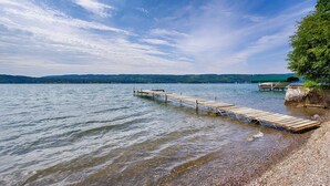 Un muelle de madera que se adentra en un lago tranquilo con una orilla rocosa y una colina boscosa a lo lejos.