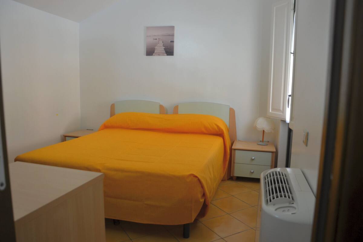 A small, neatly arranged bedroom with a bed, bedside table, lamp, and a heater.