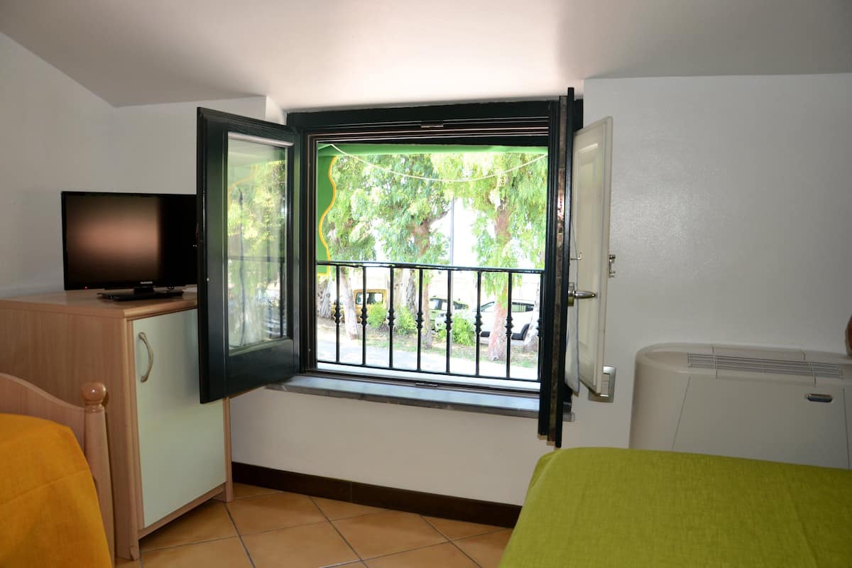 A room with a bed, a TV on a cabinet, a window with a view of trees and parked cars, and a white air conditioner.