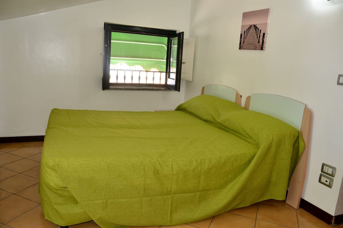 A single bed with a green bedspread in a room with white walls, a window with a view of a green field, and a small picture on the wall.