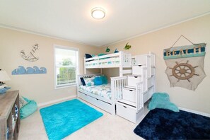 Une chambre d’enfant avec des lits superposés, une fenêtre avec des stores, une décoration murale représentant un poisson et un élément décoratif suspendu sur le thème de la plage.