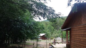 A rustic wooden cabin with a porch, surrounded by dense greenery and a dirt path leading to another structure in the background. - House with terrace near the lake, national parks and caburgua eyes (Pucon)