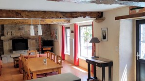 Un interior rústico con vigas de madera a la vista, una chimenea de piedra, una mesa de comedor de madera con sillas, un televisor y un amplio ventanal con cortinas rojas.