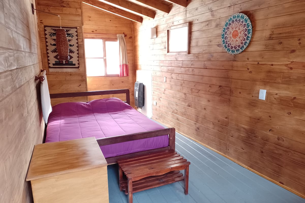 Ein Schlafzimmer einer Holzhütte mit Bett, einem kleinen Holztisch, einer Bank, einem Fenster und einer Wanddekoration.