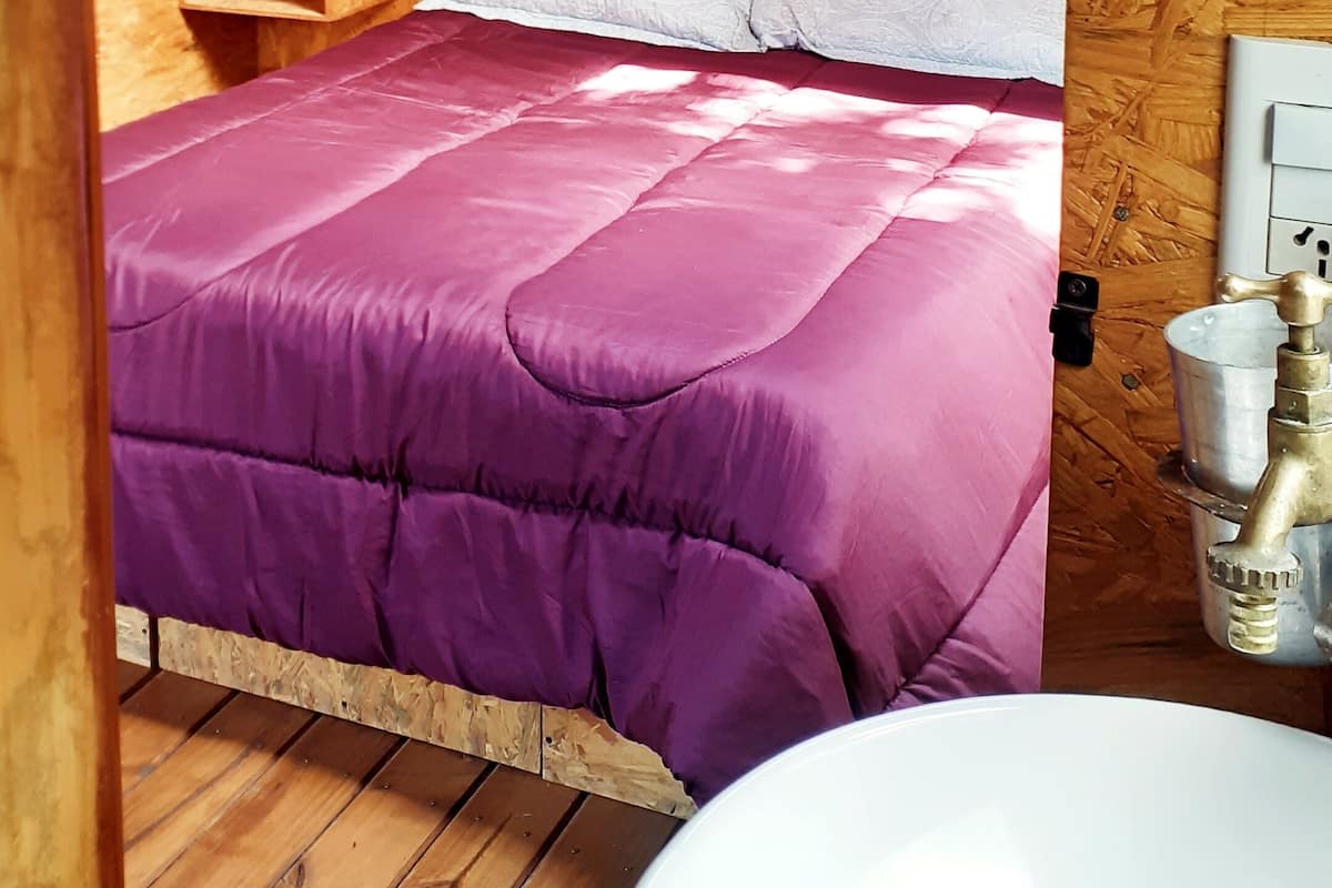 Ein Zimmer aus Holz mit einem Bett, einem Nachttisch, einer Lampe, einem Fenster und einem Waschbecken.