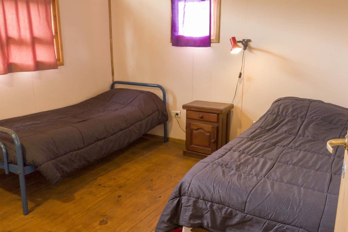 Ein kleines Zimmer mit zwei Einzelbetten, einem Holzkapfen, einem Nachttisch mit Lampe und einem Fenster mit Vorhängen.