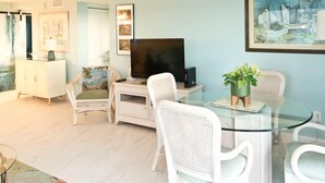 A living room with a glass coffee table, white wicker chairs, a flat-screen TV, and a painting on the wall.