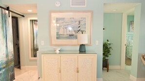 A hallway with a light blue wall, a framed painting of sailboats, a white cabinet with a glass top, and a potted plant.