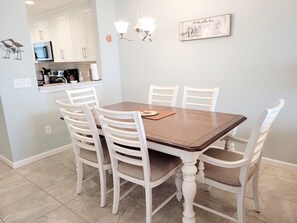 Uma sala de jantar com mesa de madeira e cadeiras brancas, um candelabro e uma cozinha ao fundo.