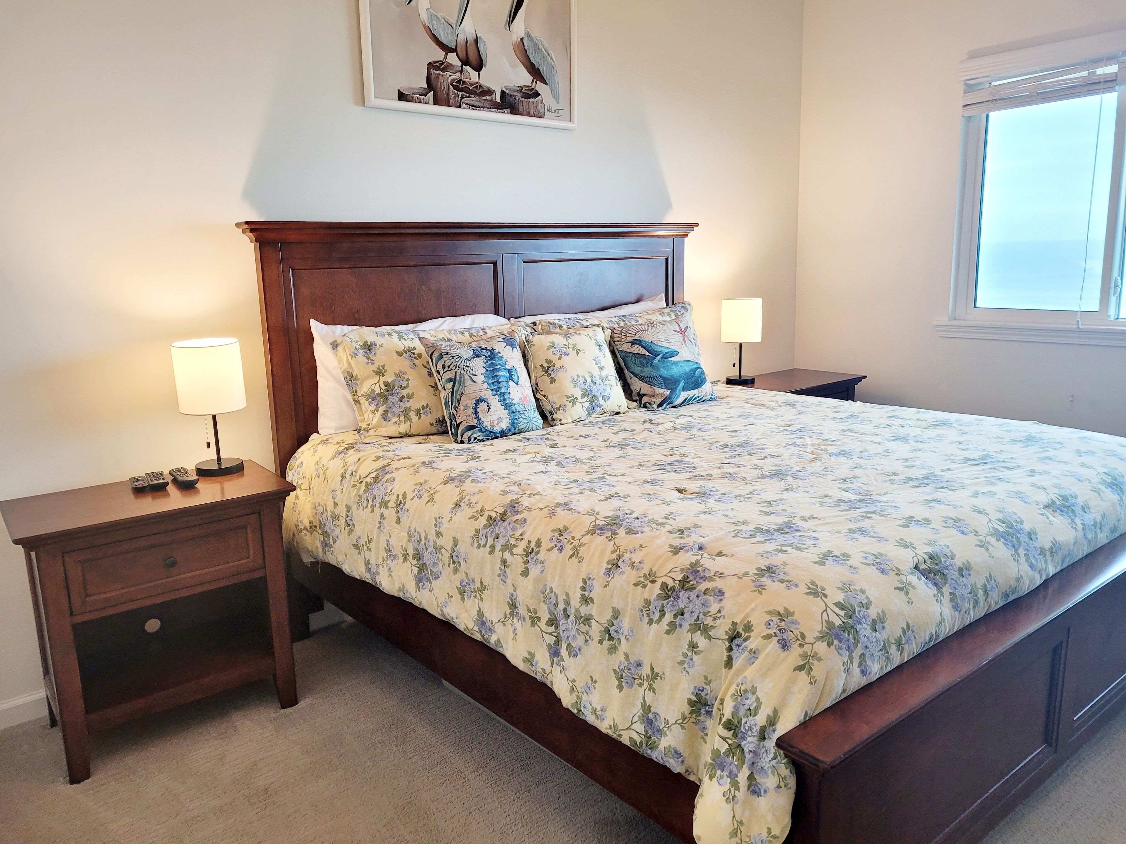 A bedroom with a large bed, two bedside tables, a nightstand, a window with blinds, and a painting on the wall.