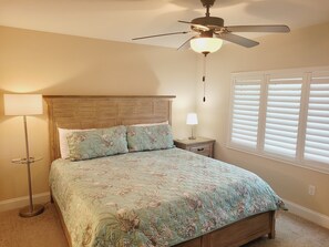 A bedroom with a bed, a ceiling fan, a nightstand with a lamp, and a window with blinds.
