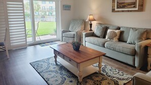A living room with a sofa, a patterned armchair, a wooden coffee table, and a lamp. There is a sliding glass door leading to a balcony with a view of a building and greenery. - Oceanview from balcony 3 Bedroom 2 Bath ground floor Unit - 110 (ST. AUGUSTINE)