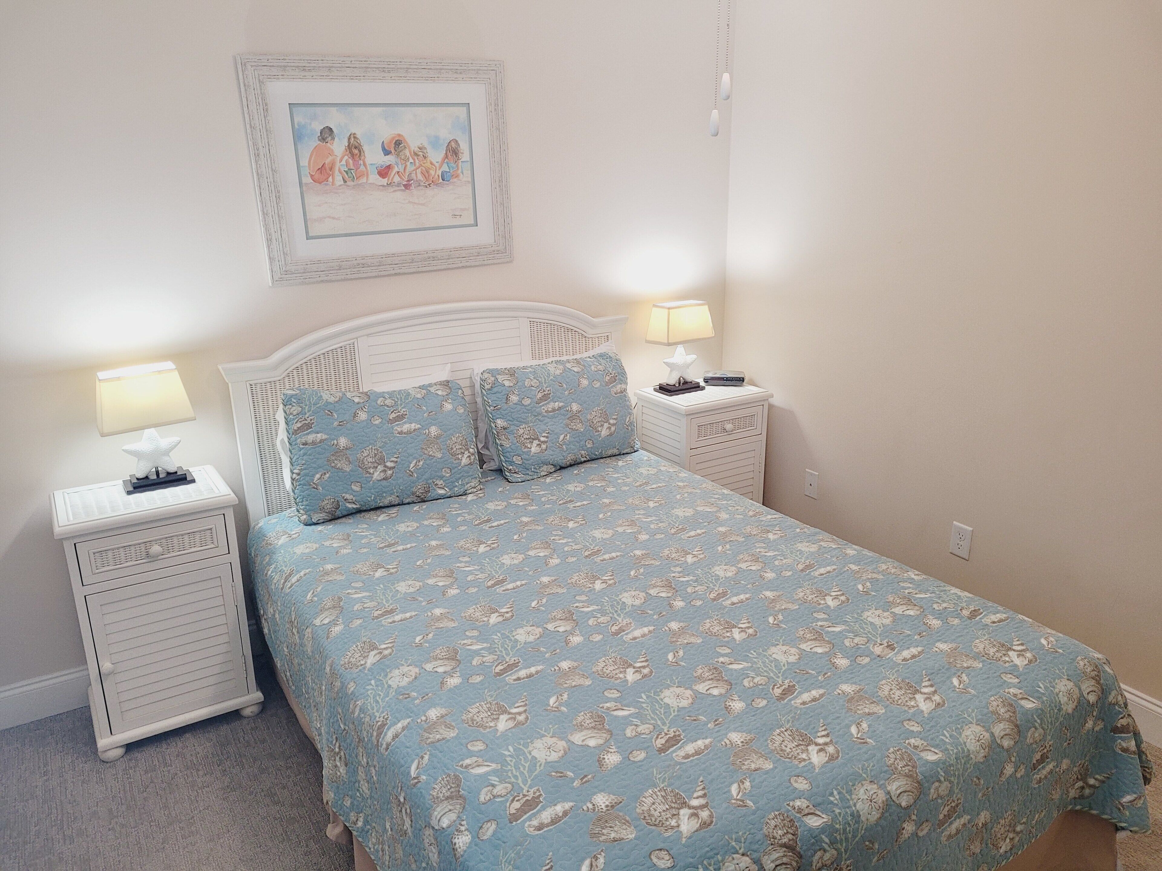 Een slaapkamer met een bed, twee nachtkastjes, een lamp op elk en een lijst aan de muur, plus een zeester decoratie op het nachtkastje.