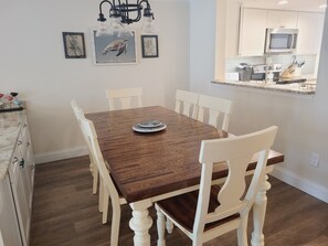 A dining area with a wooden table and white chairs, a chandelier, framed artwork, and a kitchen visible through an open doorway.