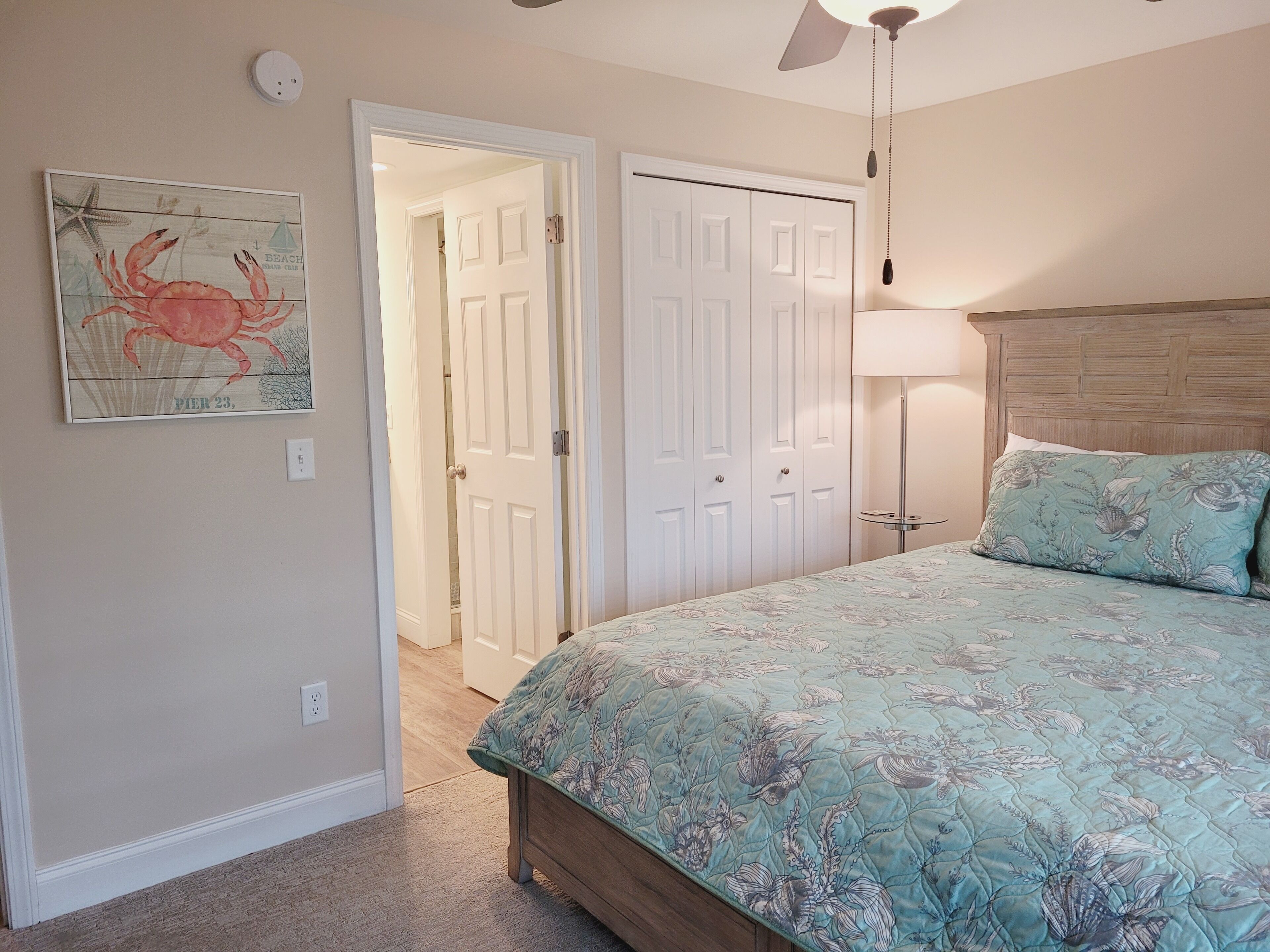 Une chambre à coucher comprenant un lit, un ventilateur de plafond, une table de chevet, une lampe et une porte donnant sur une autre pièce.
