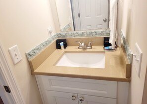 A bathroom with a beige countertop, a white sink, and a mirror above it. There are two towel racks, one with a black towel and one with a white towel.