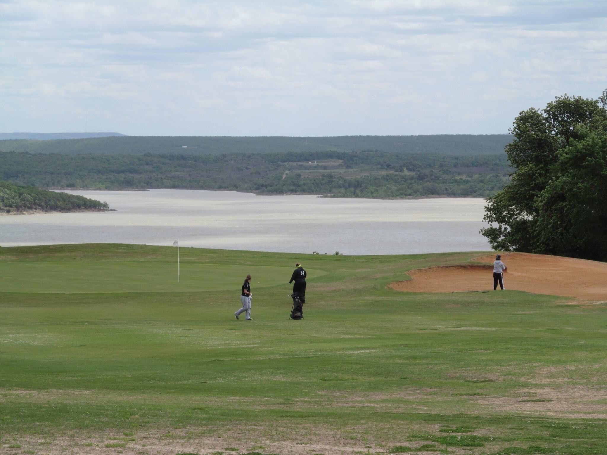 Campo de golf con una masa de agua al fondo, una bandera y un árbol.