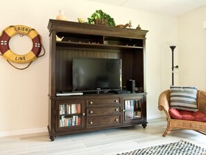 A living room with a dark wooden entertainment center, a flat-screen TV, and a wicker chair with cushions. - Fully furnished Oceanview 2 Bedroom Flat _ Unit 119 (ST. AUGUSTINE)