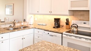 Una cocina moderna con armarios blancos, encimeras de granito, cafetera, hervidor y lavavajillas.
