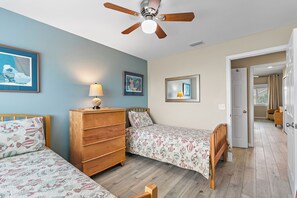 A bedroom with two single beds, a wooden dresser, a ceiling fan, and a door leading to another room.