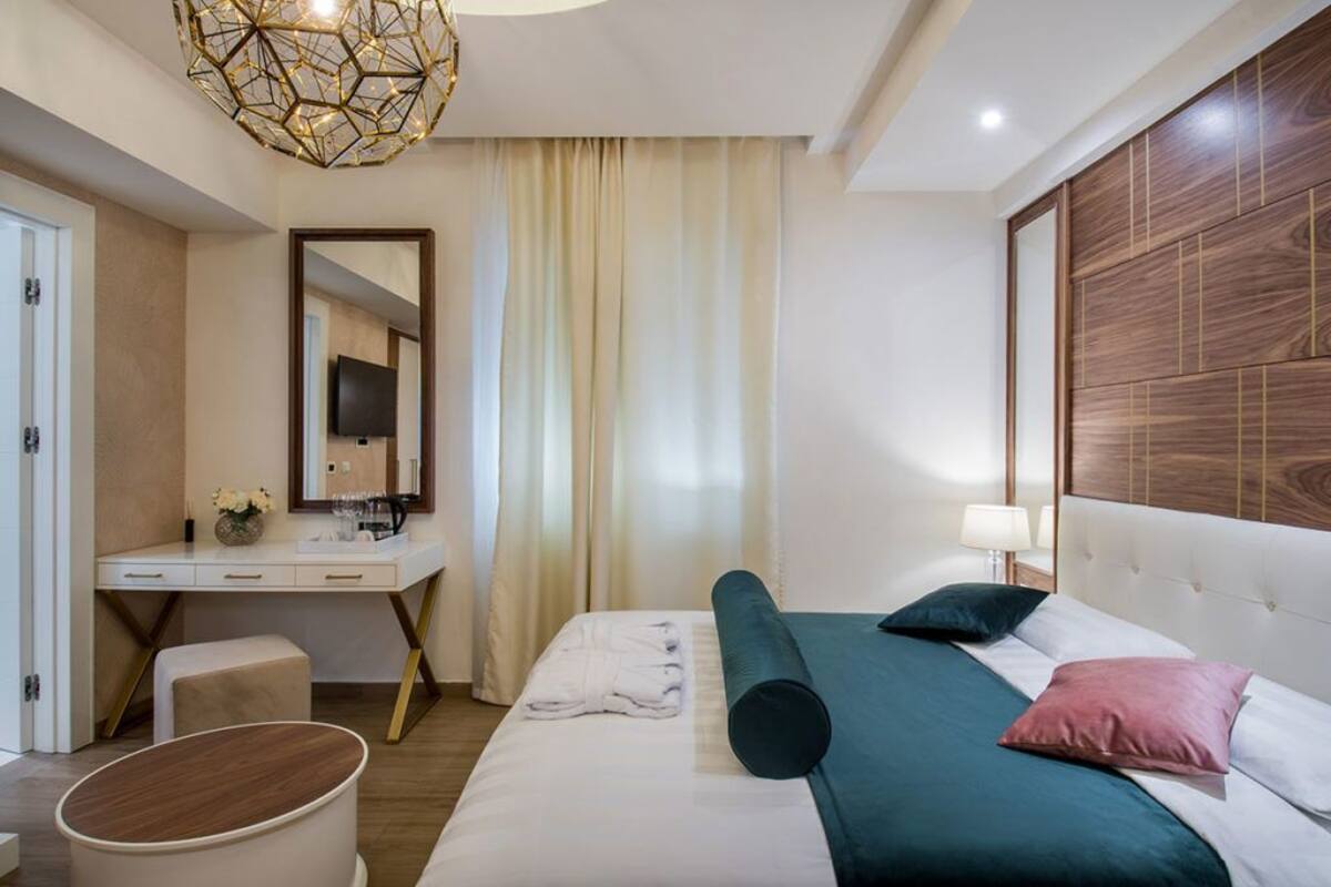 A modern hotel room with a neatly made bed, a bedside table with a lamp, a large mirror, and a wooden headboard.