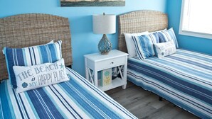 Una camera con due letti, ognuno con biancheria a righe e cuscini coordinati, un comodino bianco con una lampada e un quadro appeso che raffigura una spiaggia.