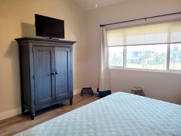 Un dormitorio con cama, armario, televisor encima, ventana con cortinas y un taburete pequeño.
