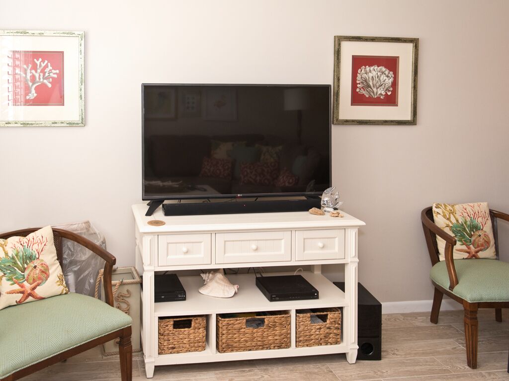 A living room with a flat-screen TV on a white TV stand, wicker baskets, and two green upholstered chairs.