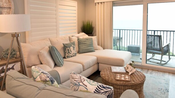 Ein modernes Wohnzimmer mit einem beigefarbenen Ecksofa, einem runden Kaffeetisch aus Rattan und einer großen Schiebeglastür, die auf einen Balkon mit Gartenmöbeln führt.