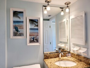 Un baño con encimera de granito, un lavabo blanco, un espejo grande y dos cuadros con escenas de playa.
