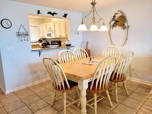 Un coin repas avec une table en bois et des chaises blanches, une cuisine en arrière-plan et un élément décoratif mural au-dessus du miroir.