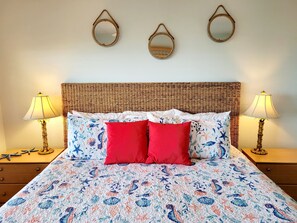 Una cama con cabecera trenzada, dos almohadas rojas y una colcha con estampado floral. Hay dos lámparas de noche y tres espejos de pared.