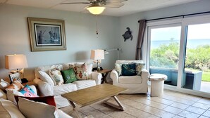 Sala de estar com sofá branco, mesa de centro de madeira e poltrona branca. Há um quadro na parede, um abajur e vista para o exterior por uma porta de vidro.