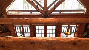 Interior de una cabaña de troncos con un techo de gran viga de madera, chimenea de piedra, banca de madera, sofá de cuero y vistas del paisaje nevado a través de amplios ventanales.