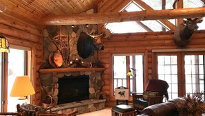 Interior de una cabaña de troncos con chimenea de piedra, muebles de cuero y un venado montado en la pared.