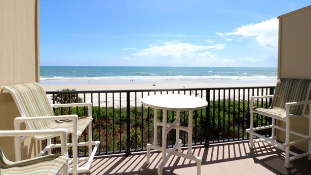 Un balcon avec des chaises blanches et une petite table, donnant sur une plage sous un ciel dégagé.