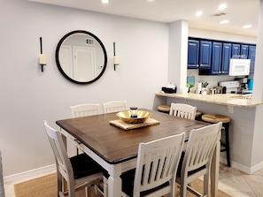 A dining area with a wooden table and white chairs, a round mirror on the wall, and a kitchen area with blue cabinets and a white microwave.