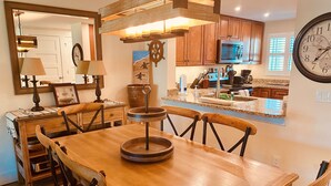 Een eethoek met een houten tafel en stoelen, een keuken met houten kasten en een magnetron, en een wandklok.