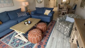 A living room with a blue sofa, a wooden coffee table, patterned ottomans, a patterned rug, and a dining area with a wooden table and chairs.