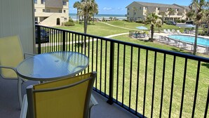 A balcony with a glass table and chairs, overlooking a swimming pool and a grassy area with palm trees.