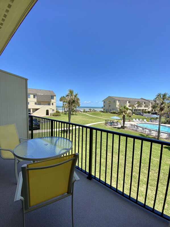 A balcony with a glass table and chairs, overlooking a swimming pool and a grassy area with palm trees.