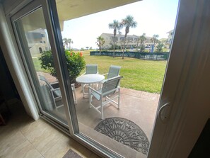Une porte vitrée donne sur un patio extérieur agrémenté d’une table ronde et de deux chaises, d’un petit tapis décoratif, et offre une vue sur un espace verdoyant avec des palmiers et un bâtiment en arrière-plan.