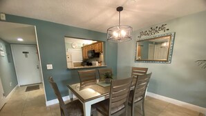 Un coin repas avec une table et des chaises, un lustre, une cuisine en arrière-plan et un miroir au mur.