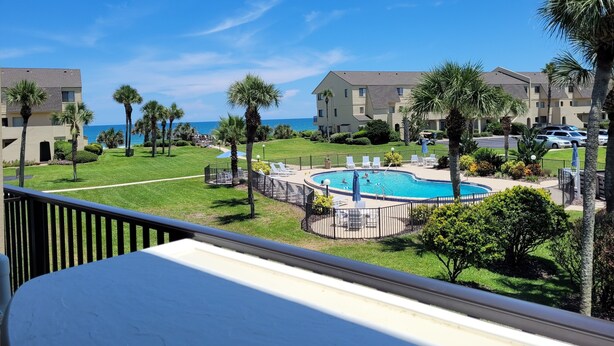 A pool area with a view of the ocean, surrounded by palm trees and a building with a flat roof.