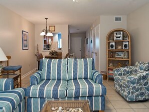 A living room with striped sofas, a coffee table with seashells, a bookshelf, and a dining area in the background.