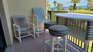 A balcony with two chairs and a small table, overlooking a grassy area with palm trees and a building in the background.