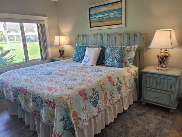 A bedroom with a large bed, two bedside tables with lamps, a painting above the bed, and a view of the outside through a window.
