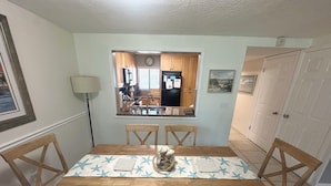Un coin repas avec une table et des chaises en bois, une fenêtre donnant sur la cuisine et un tableau encadré au mur.