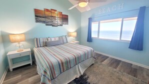 Une chambre à coucher comprenant un lit, des tables de chevet, un ventilateur de plafond, une décoration murale sur le thème de la plage, une fenêtre donnant sur l’océan et un tapis.