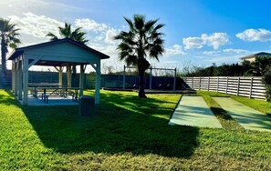 Relax in a serene outdoor setting with a shaded pavilion, bench, and table, surrounded by a well-maintained lawn and a palm tree.